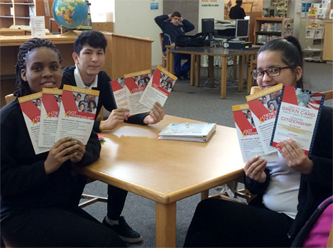 Three People sitting at a table in the library holding up citizenship information cards.