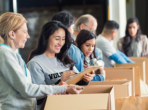 Picture of a line of people standing behind boxes and a woman is looking in one box while holding a clipboard.